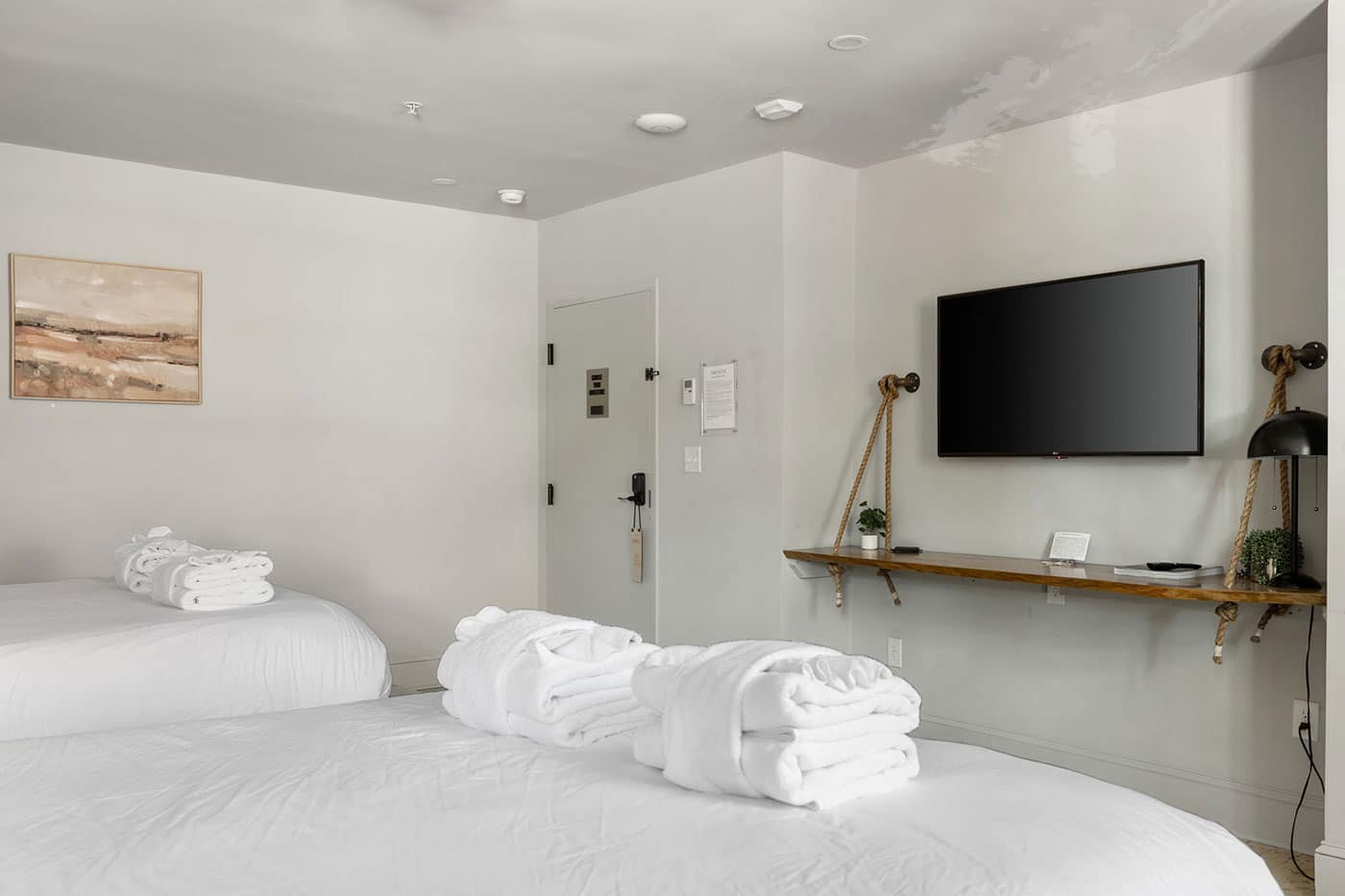 Hotel room with two white beds, neatly folded towels, and a wall-mounted flat-screen TV on a rope-supported shelf.