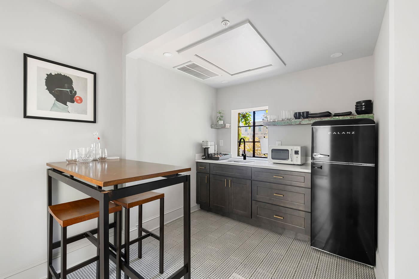 Modern compact kitchen with dark gray cabinetry, a black Galanz refrigerator, microwave, and a small high wooden table with stools; artwork on the wall and a window above the sink area.