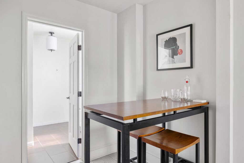 Minimalist dining nook with a wooden high table, two matching stools, and a few glassware items on the table; doorway visible to the left.