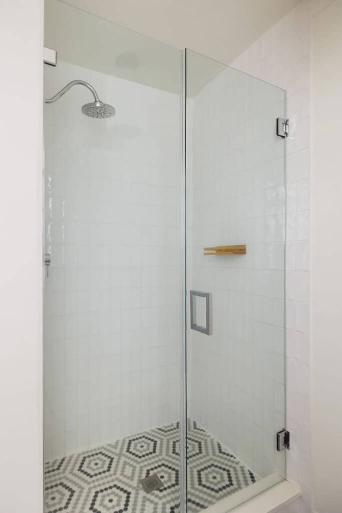 Glass-walled shower enclosure with rainfall showerhead, white tile walls, and a black-and-white hex tile floor in a modern bathroom.