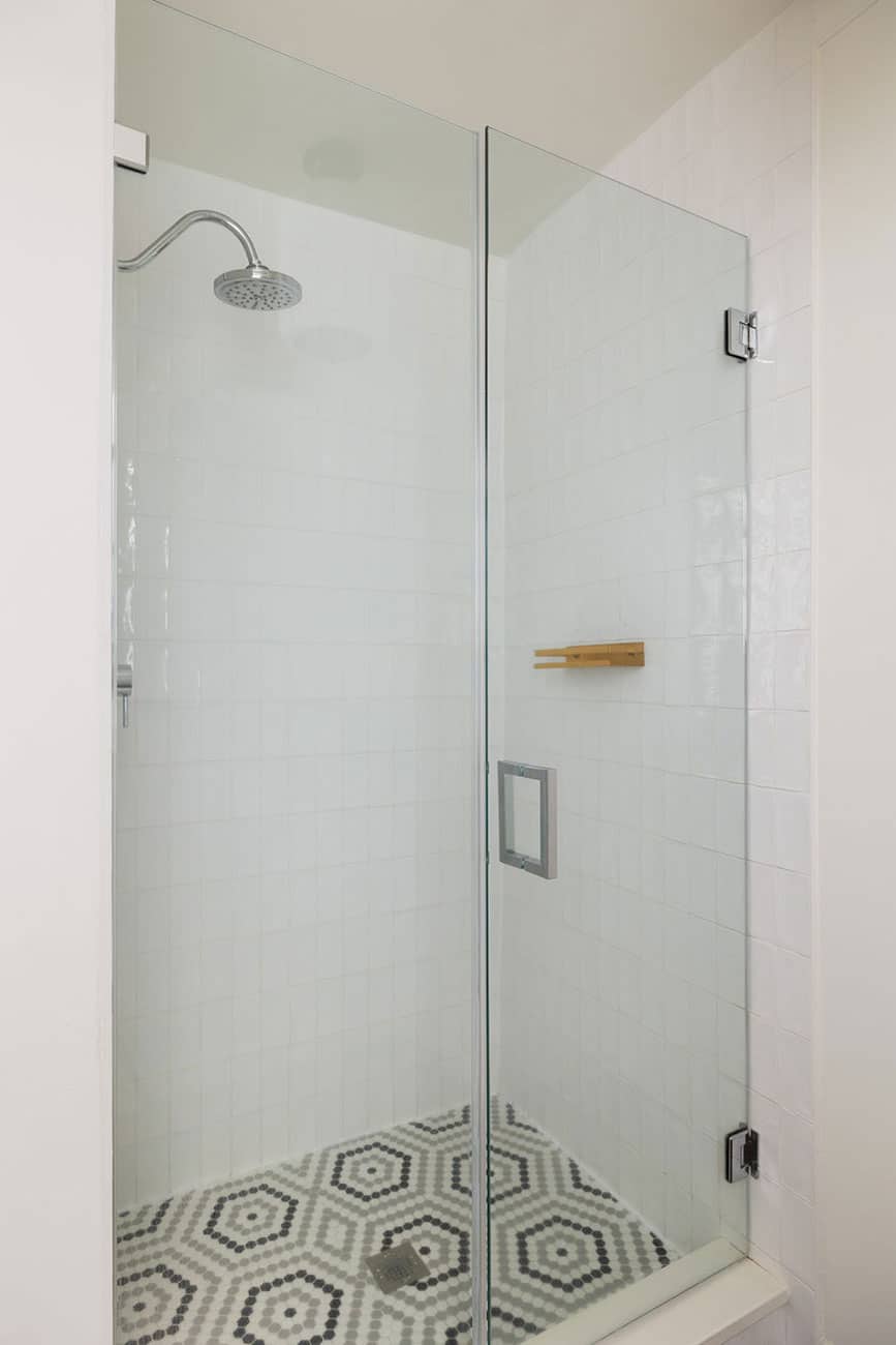 Glass-walled shower enclosure with rainfall showerhead, white tile walls, and a black-and-white hex tile floor in a modern bathroom.