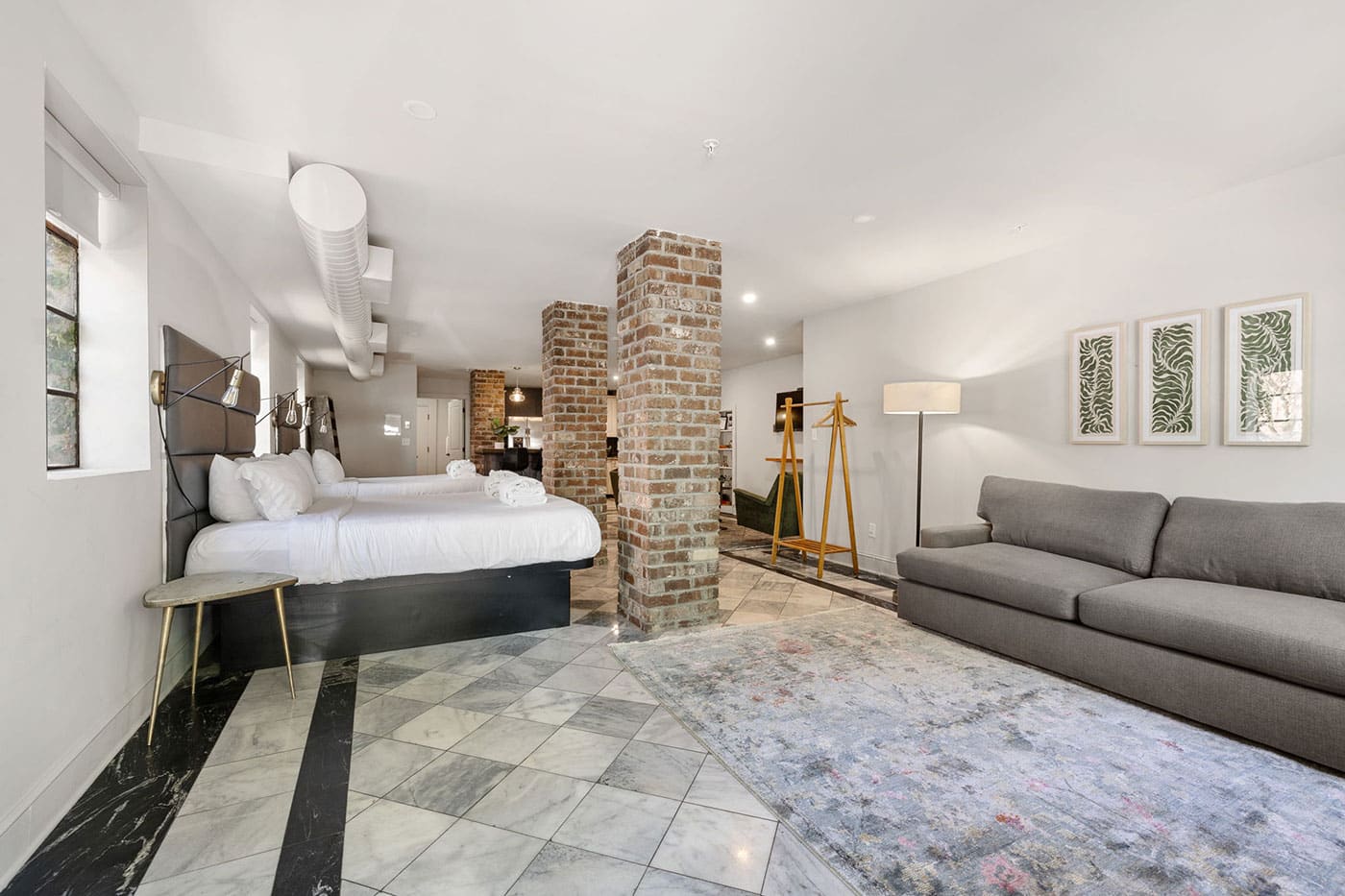 Industrial hotel suite with two queen beds, brick columns, a gray sofa, and a gold-legged side table.