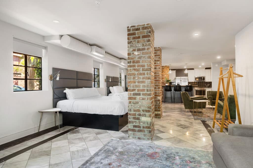 Two queen beds with padded headboards in a modern open-plan suite, brick columns, marble floors, and a visible kitchen with bar stools in the background.