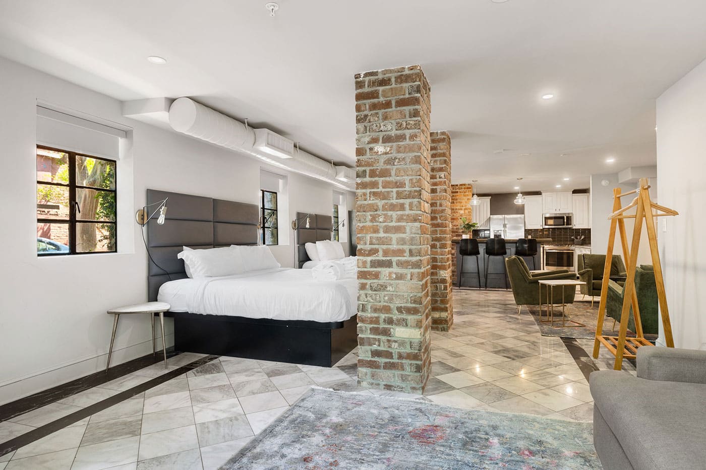 Two queen beds with padded headboards in a modern open-plan suite, brick columns, marble floors, and a visible kitchen with bar stools in the background.