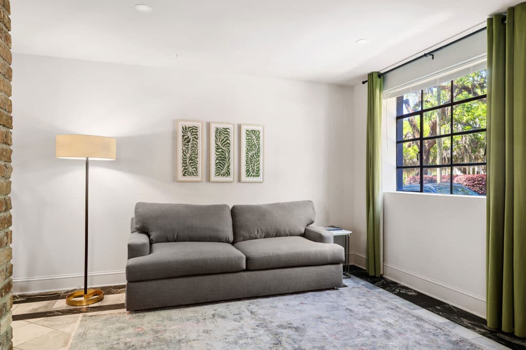 Living room with a gray sofa, a tall floor lamp, and three framed fern prints on a white wall; green curtains frame a large window.