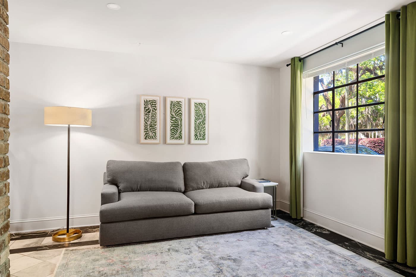 Living room with a gray sofa, a tall floor lamp, and three framed fern prints on a white wall; green curtains frame a large window.