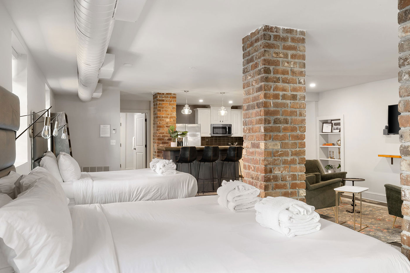 Spacious loft suite with two white beds, brick columns, open kitchen, and living area with chairs.