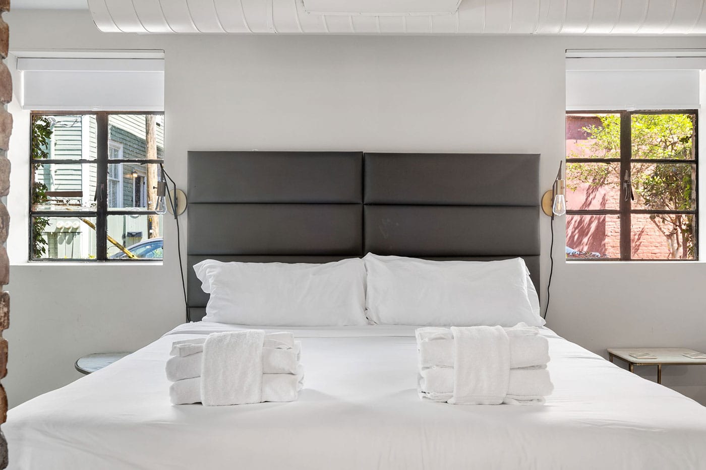 Bedroom with a large gray padded headboard, white bedding, and two rolled towels on the bed. Windows let in natural light.
