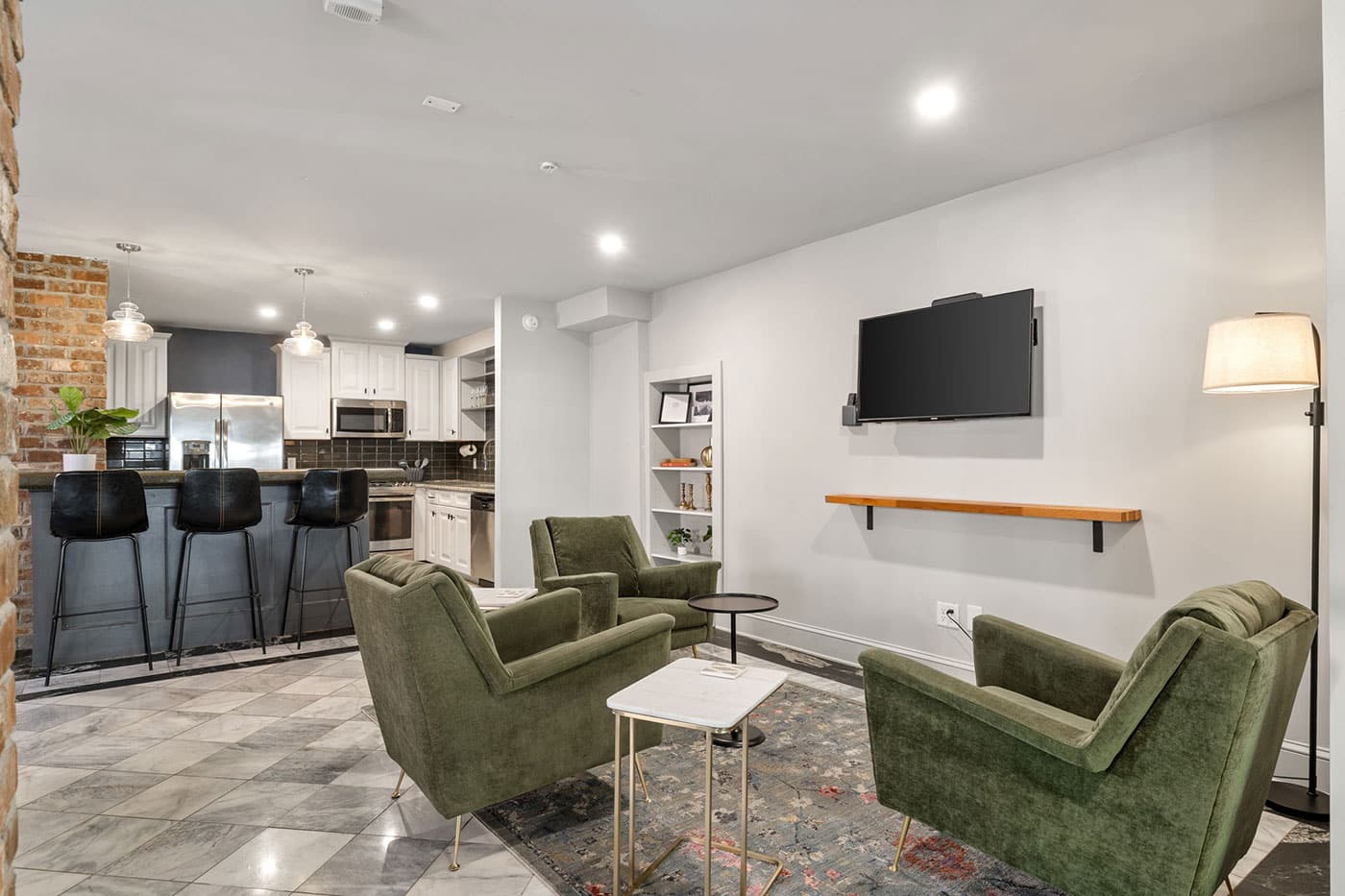 Open-concept living area with green armchairs, a TV on the wall, and a floating wooden shelf above a small table lamp.