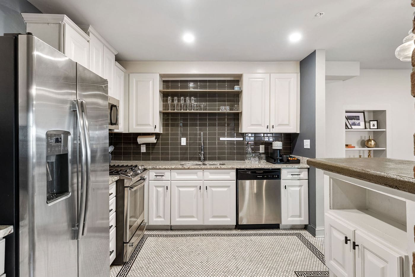 Modern kitchen with stainless refrigerator, oven, and dishwasher, white cabinets, and a dark gray tile backsplash above a granite counter.