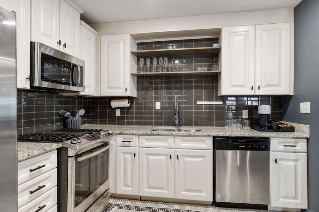Modern L-shaped kitchen with white cabinets, stainless appliances, and a dark tile backsplash; open shelves hold glassware above the sink area.