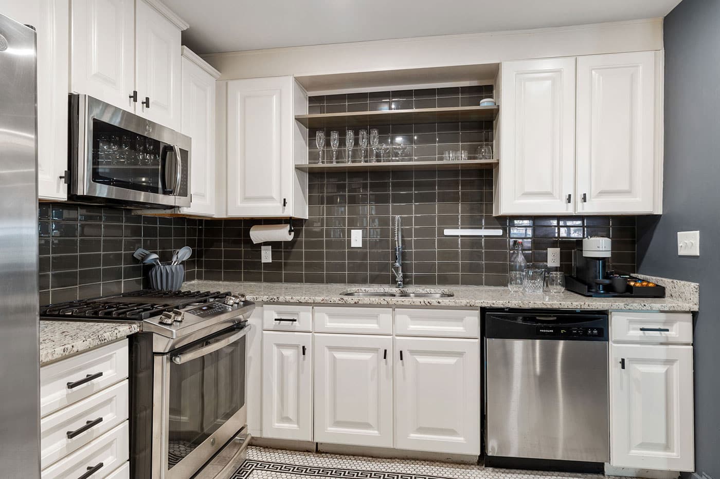 Modern L-shaped kitchen with white cabinets, stainless appliances, and a dark tile backsplash; open shelves hold glassware above the sink area.