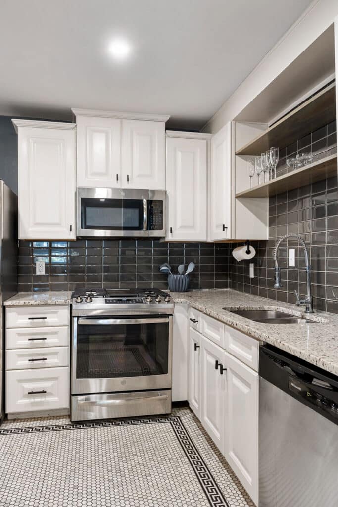 Modern kitchen with white cabinets, stainless steel stove and microwave, dark glass tile backsplash, open shelving with glassware, and a speckled countertop.
