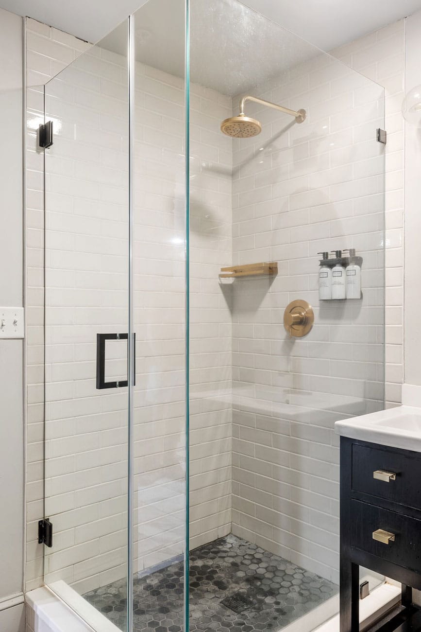 Glass-enclosed shower with white subway tiles, brass rain showerhead, and wall-mounted shampoo dispensers; dark vanity nearby.