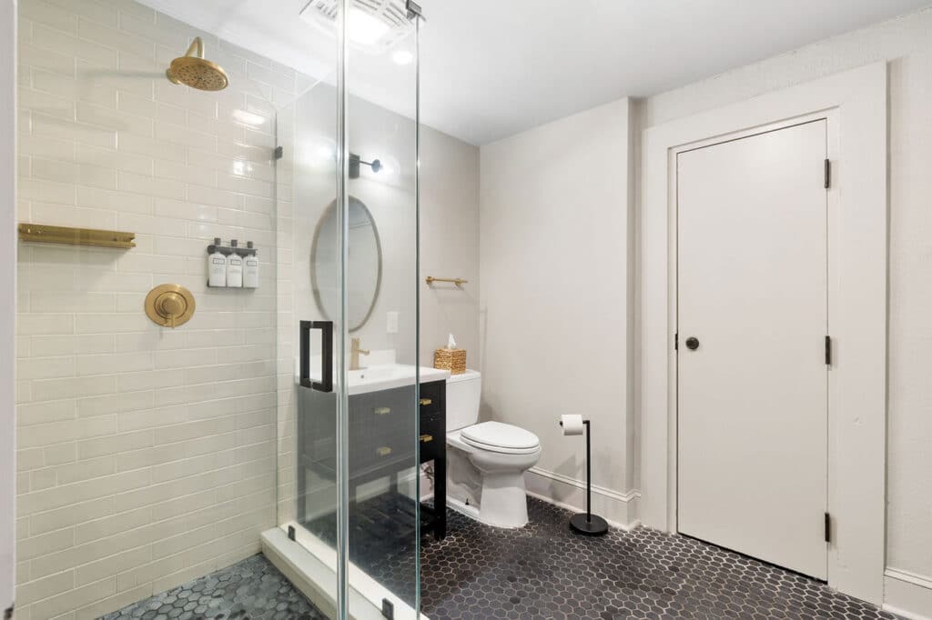 Modern bathroom with a glass-enclosed shower, white subway tile, and brass fixtures, adjacent to a white toilet and black vanity against pale walls.
