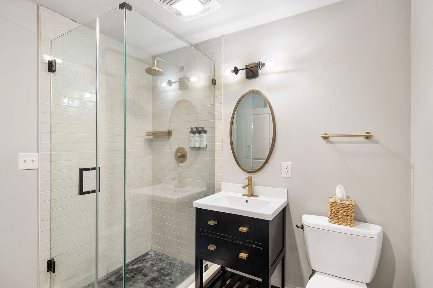 Bathroom with glass-enclosed shower, white subway tiles, and a black vanity featuring brass hardware and a white sink. Oval mirror above, brass faucet, and a tissue basket on the toilet tank are visible.