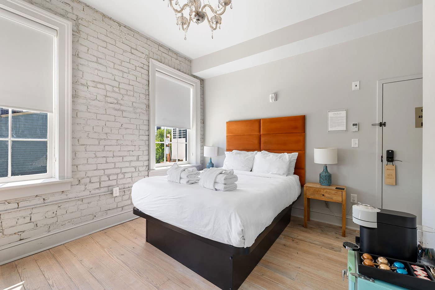 Bright hotel-style bedroom with white bedding, orange padded headboard, and two windows with white blinds opposite a brick wall interior.