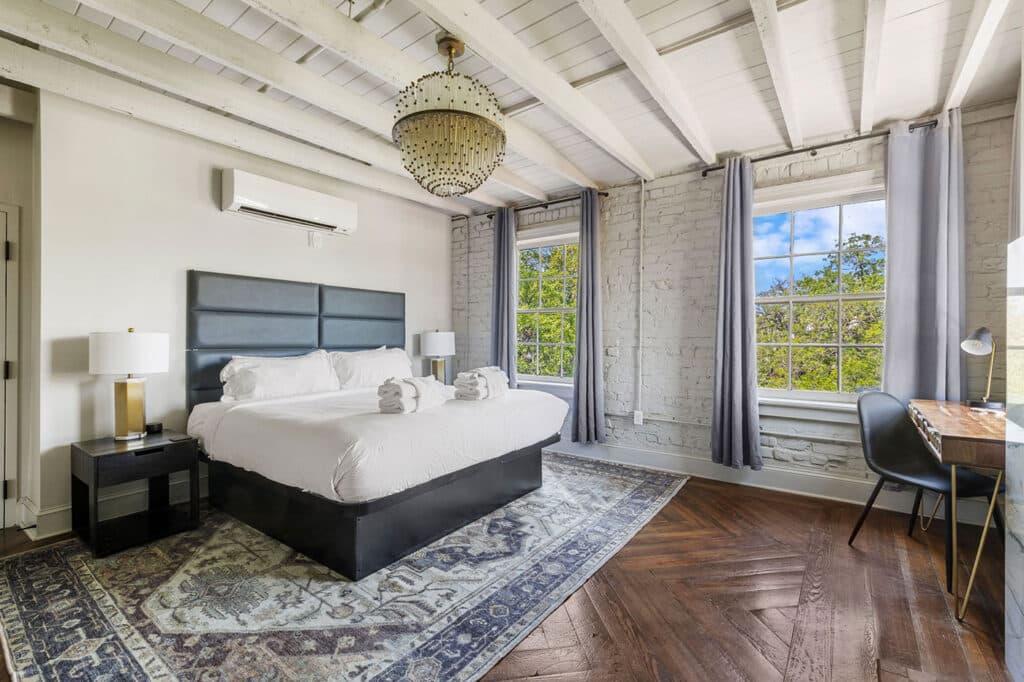 Bright industrial bedroom with white brick walls, exposed beams, a king bed with gray headboard, two nightstands, chandelier, and a rug.
