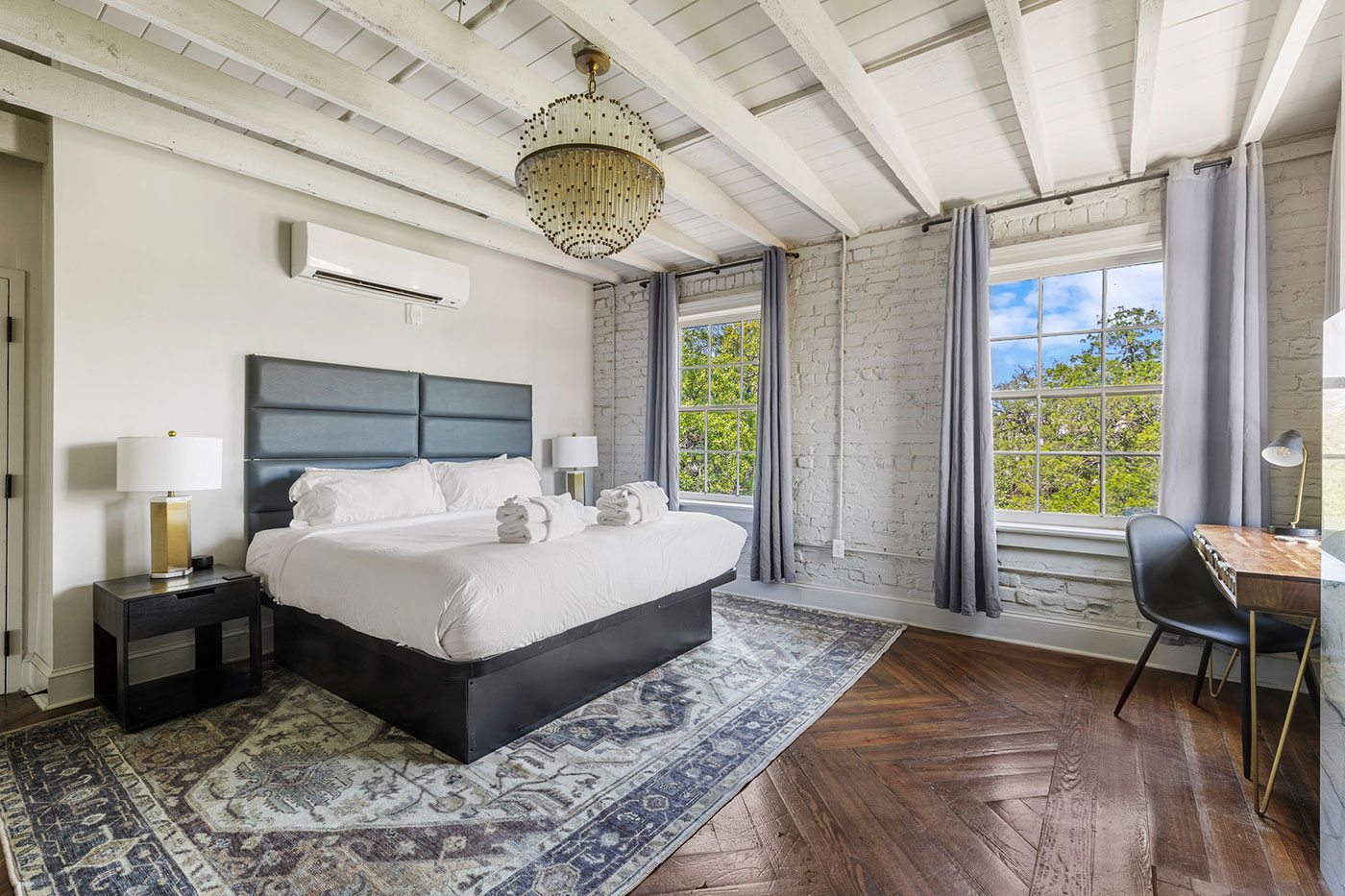 Bright industrial bedroom with white brick walls, exposed beams, a king bed with gray headboard, two nightstands, chandelier, and a rug.