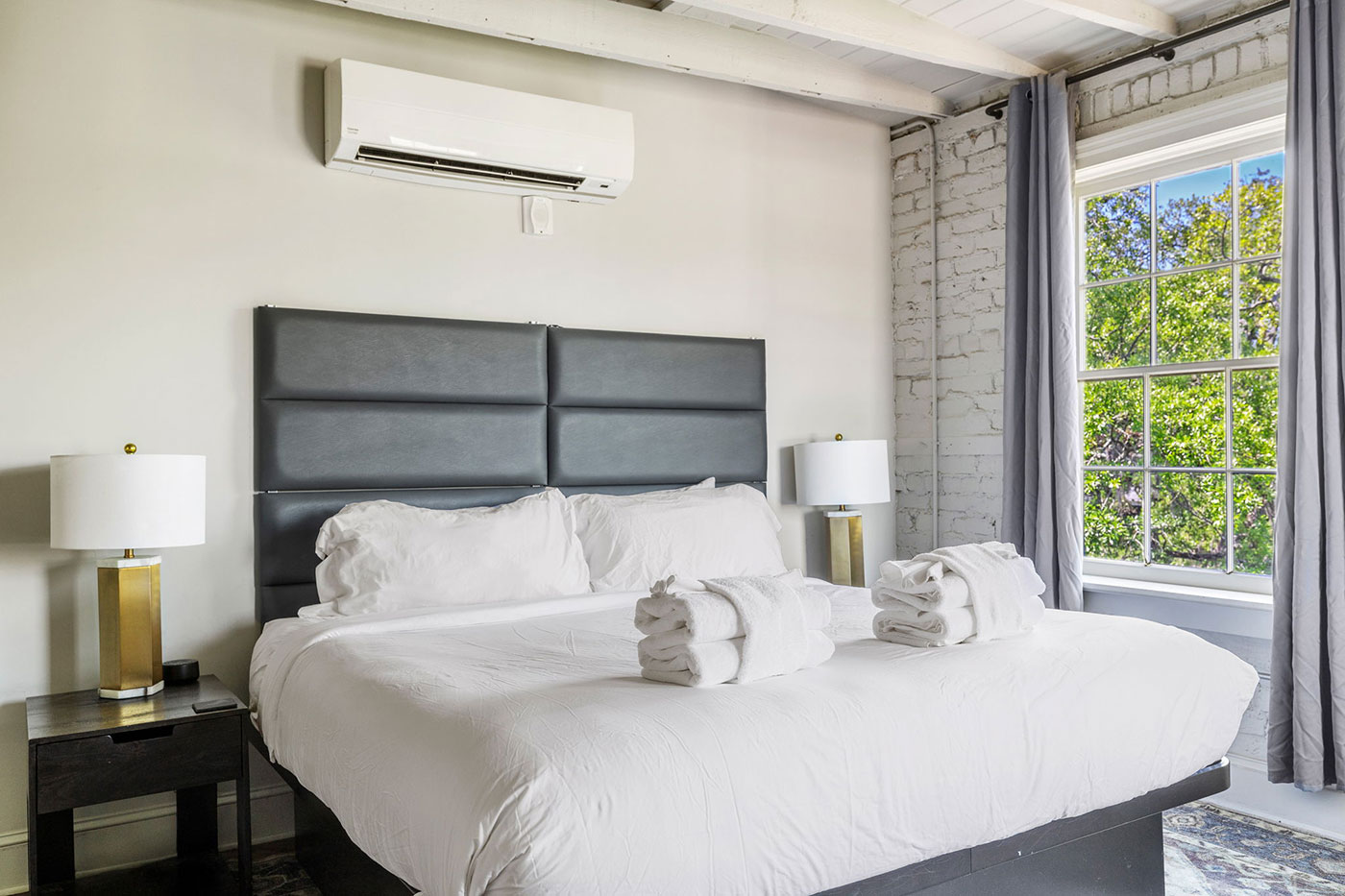 Modern bedroom with a king bed, white linens, and a padded gray headboard, two folded towels on the bed, and gold lamps on dark nightstands on either side.