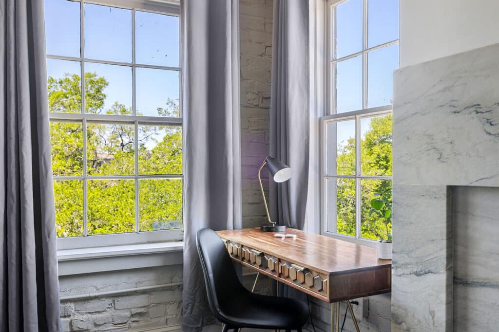 A wooden desk and black chair by a large window with gray curtains and a modern lamp, overlooking green trees outside.