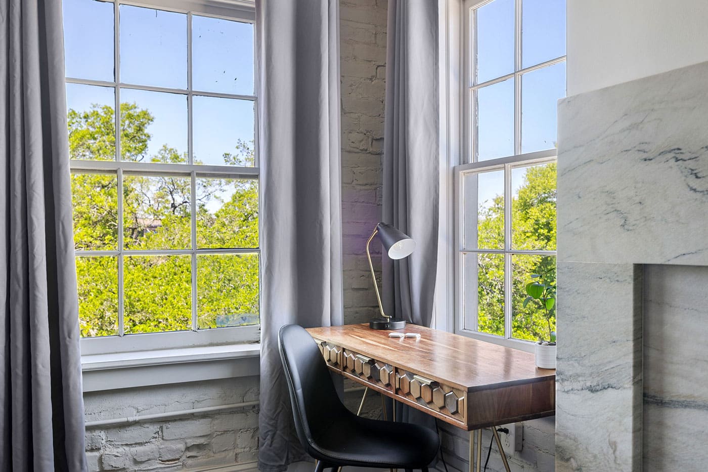 A wooden desk and black chair by a large window with gray curtains and a modern lamp, overlooking green trees outside.
