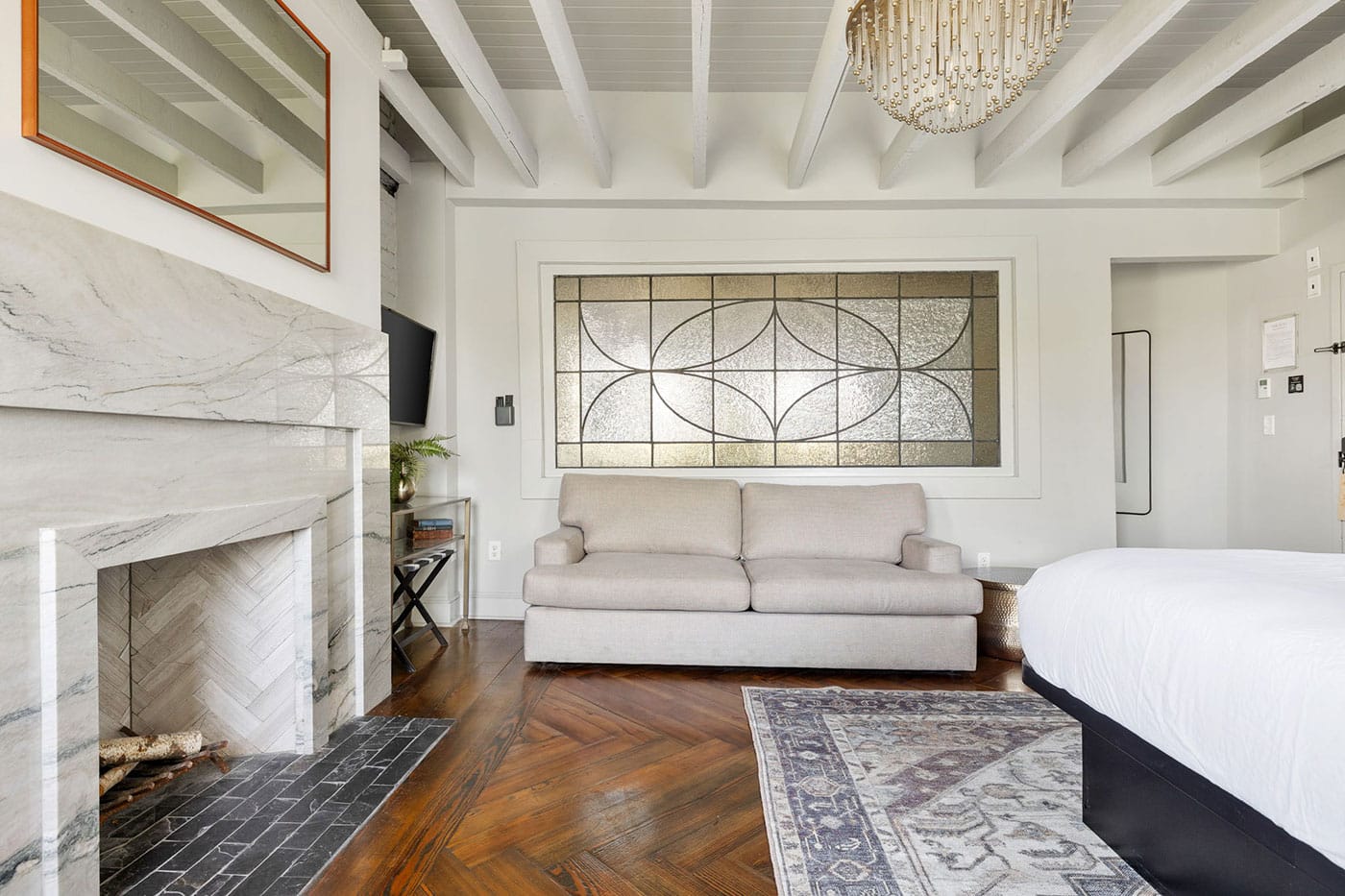Modern hotel-style suite: marble fireplace, beige sofa, decorative stained-glass window, chandelier, and a bed in the foreground.