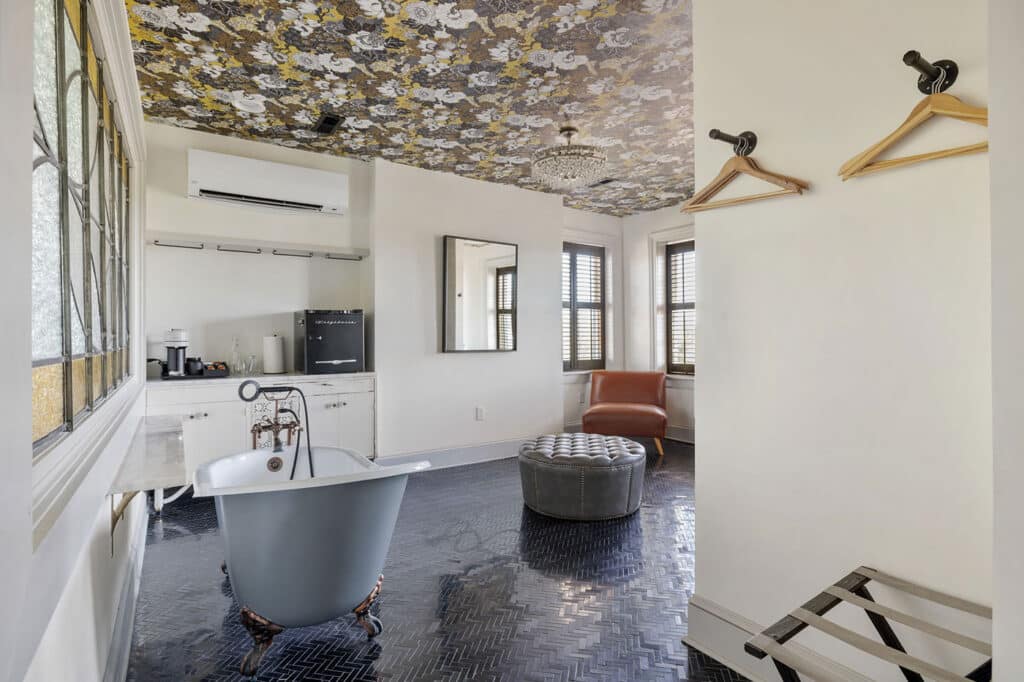 Bathroom with a gray freestanding clawfoot tub on a black herringbone floor, tufted ottoman, and brown leather chair by windows with shutters; floral ceiling pattern above.