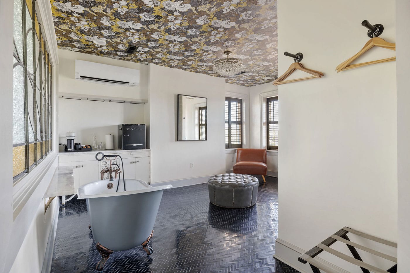 Bathroom with a gray freestanding clawfoot tub on a black herringbone floor, tufted ottoman, and brown leather chair by windows with shutters; floral ceiling pattern above.