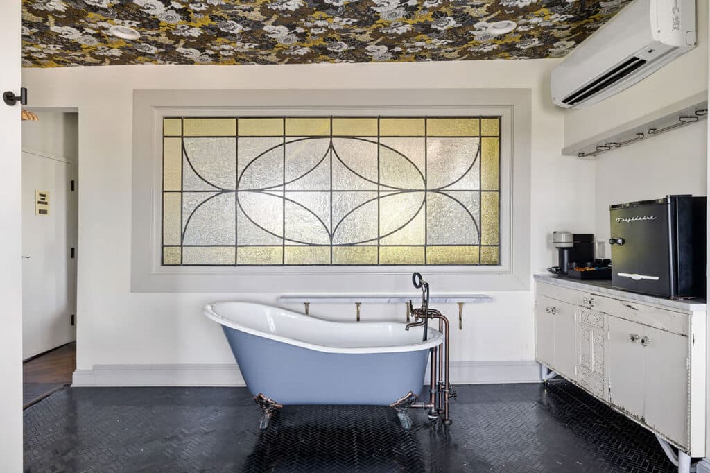Blue freestanding clawfoot bathtub with copper fixtures in a vintage bathroom, large stained glass window behind it.