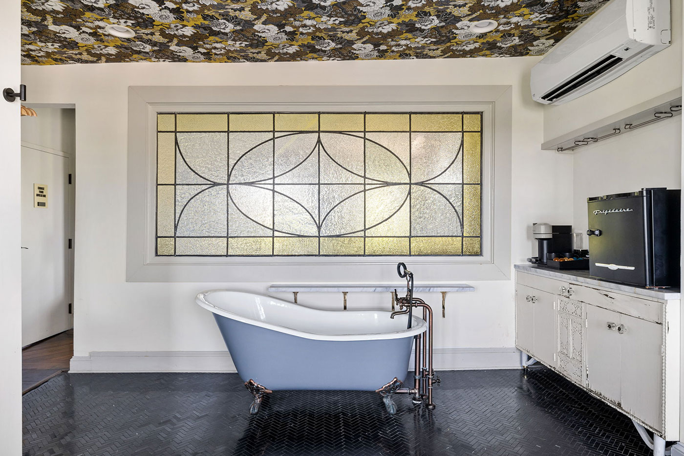 Blue freestanding clawfoot bathtub with copper fixtures in a vintage bathroom, large stained glass window behind it.