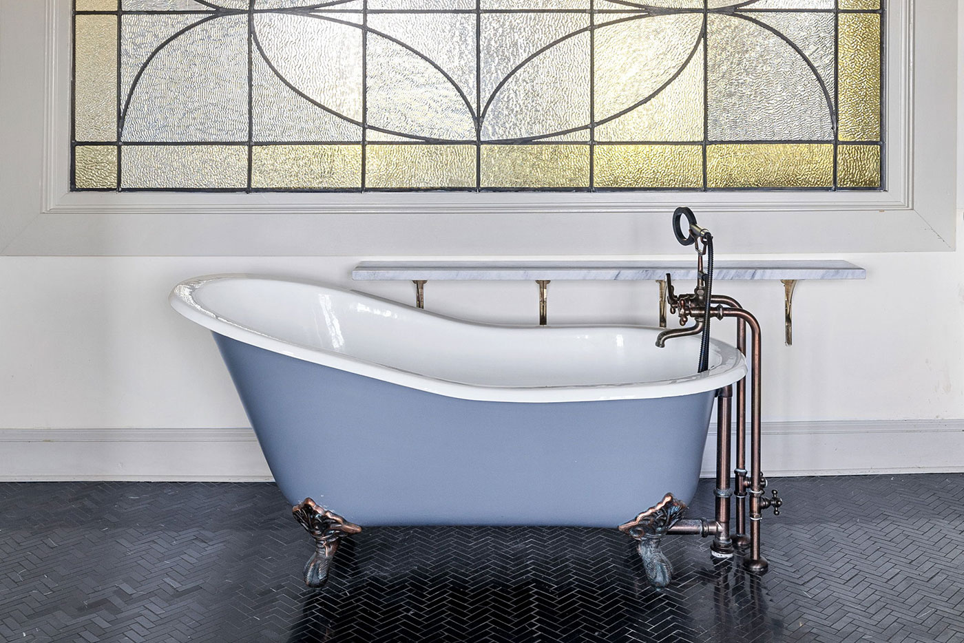 Blue freestanding clawfoot tub with white interior and copper pipes in a bathroom with a stained-glass window outside or above it.