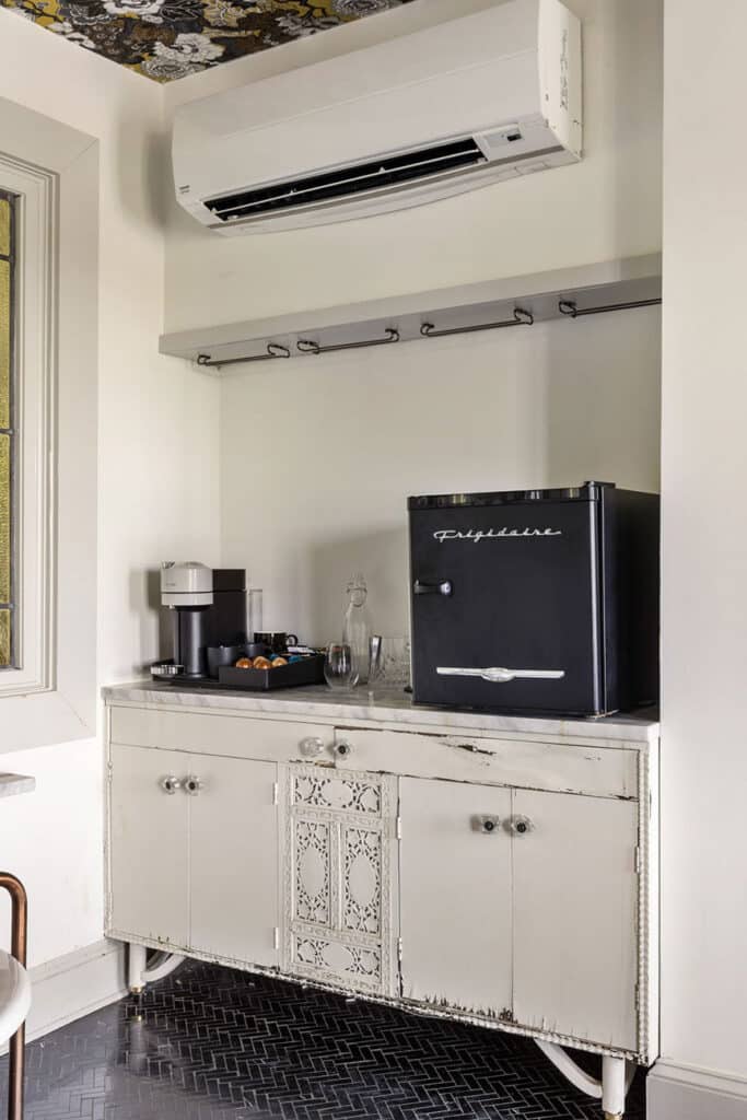 Vintage-style kitchenette with a white cabinet, a black Frigidaire mini-fridge, coffee station, and glassware on the counter under a wall-mounted air conditioner.
