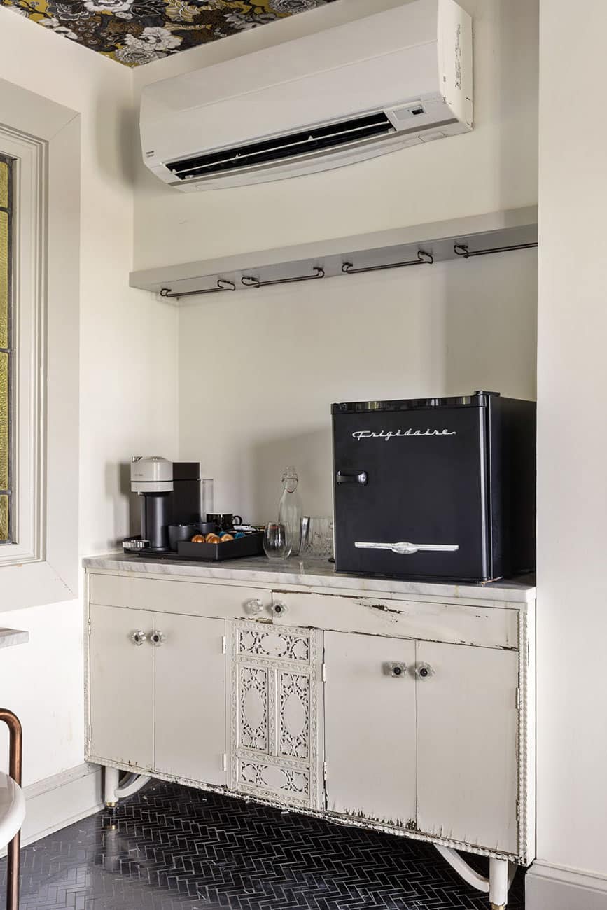 Vintage-style kitchenette with a white cabinet, a black Frigidaire mini-fridge, coffee station, and glassware on the counter under a wall-mounted air conditioner.