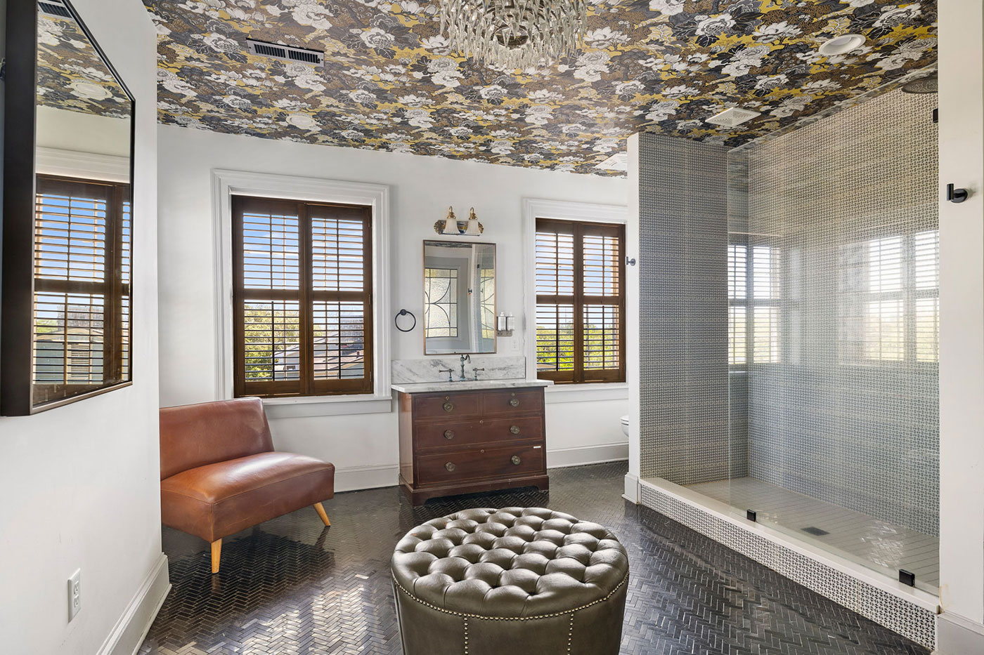 Luxurious bathroom with tufted ottoman, leather chair, marble-topped vanity, glass shower, two shuttered windows, and a floral ceiling.