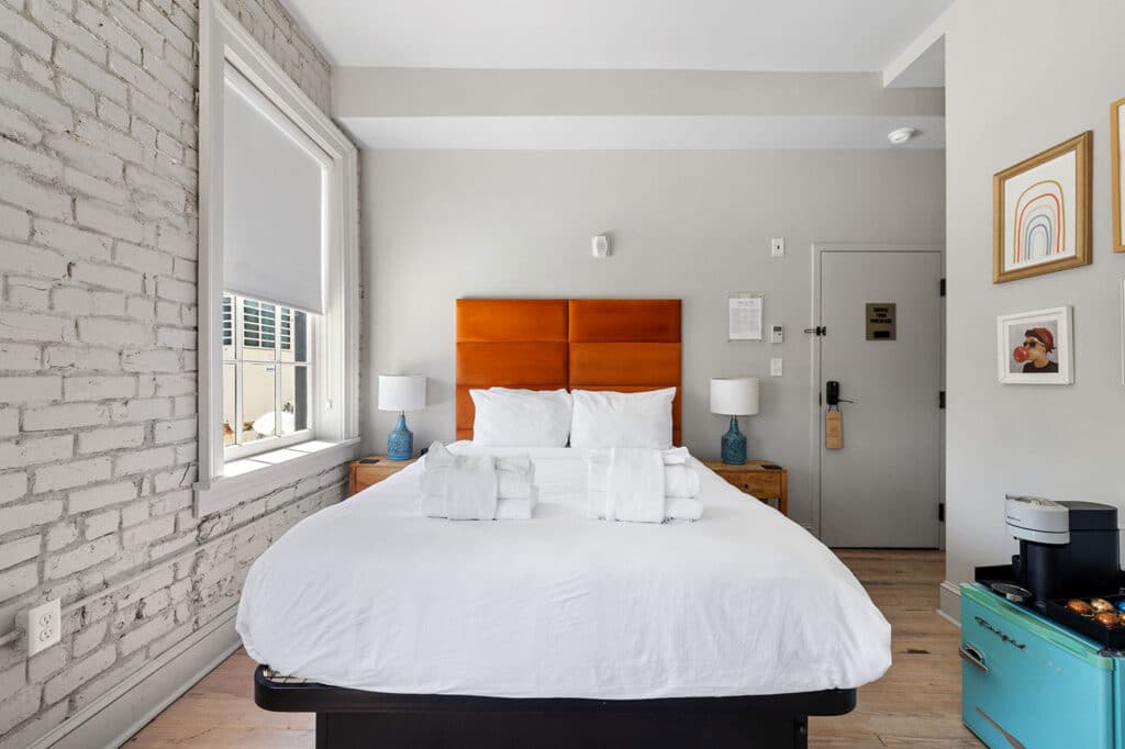 Bright hotel bedroom with an orange padded headboard, white bedding, and blue lamps on wooden nightstands under a large window with blinds.