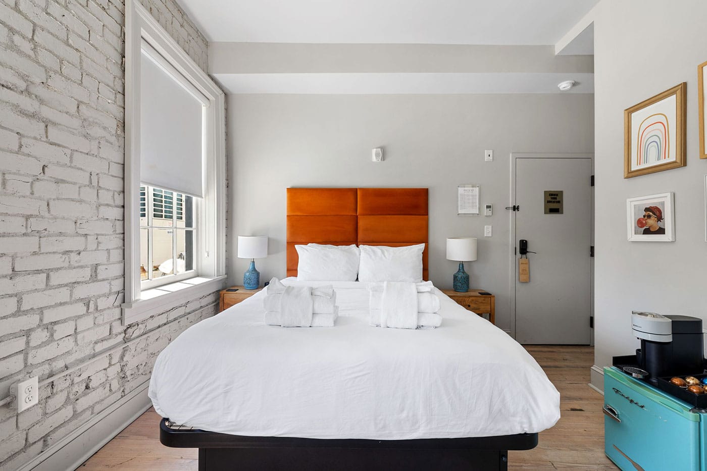 Bright hotel bedroom with an orange padded headboard, white bedding, and blue lamps on wooden nightstands under a large window with blinds.