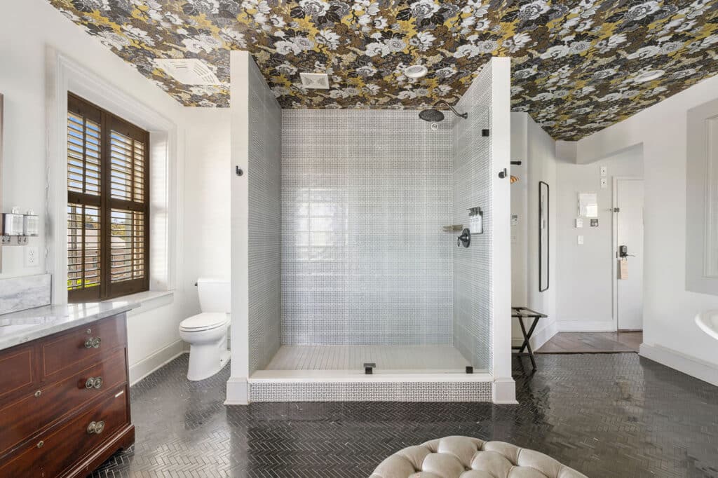 Bright bathroom with a walk-in shower, dark herringbone floor, wooden vanity, and a bold floral ceiling pattern.