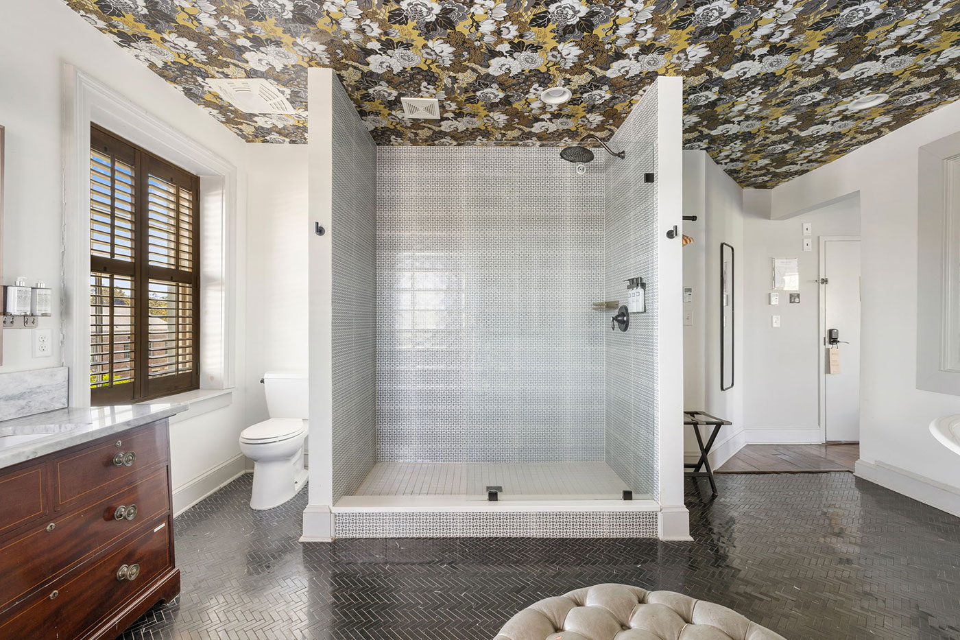 Bright bathroom with a walk-in shower, dark herringbone floor, wooden vanity, and a bold floral ceiling pattern.