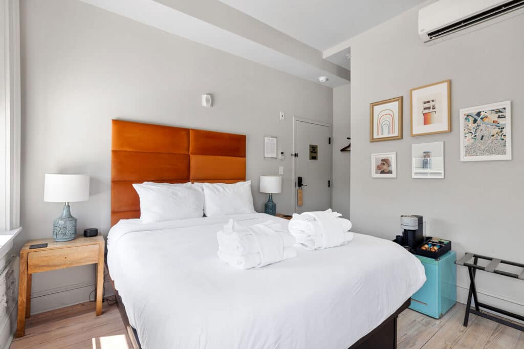 Hotel bedroom with orange padded headboard, white bedding, and rolled towels on the bed in a bright room.