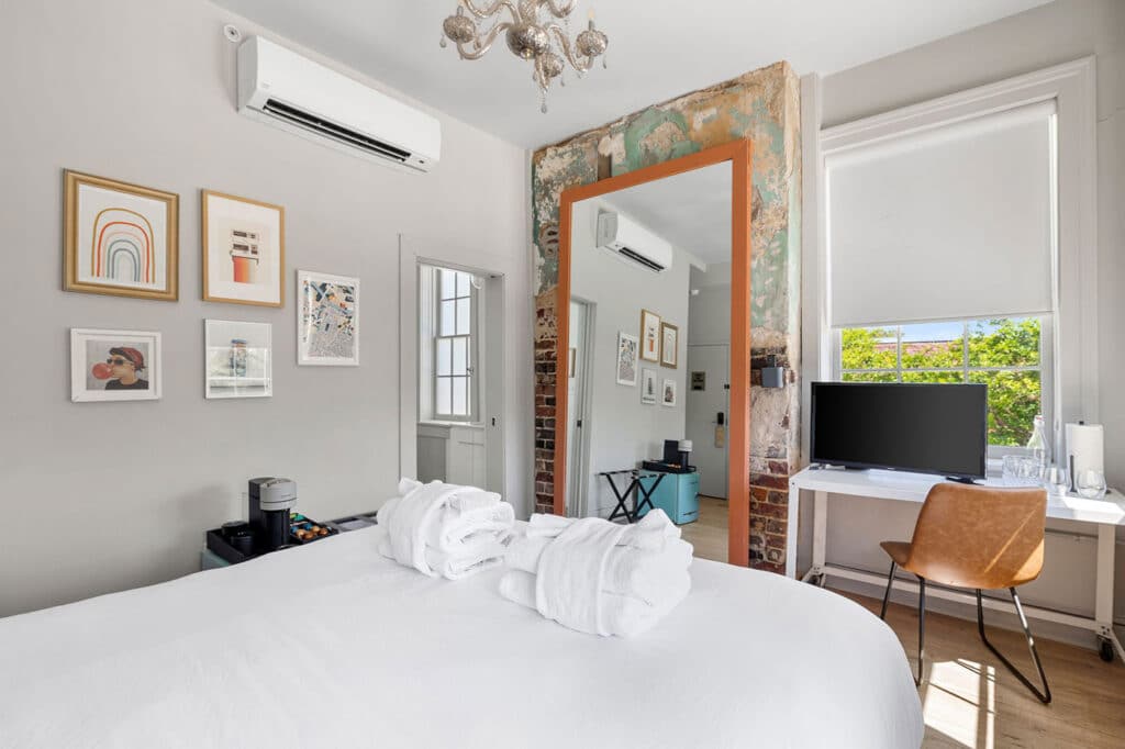 Bright bedroom with a white bed and two fluffy robes, wall art, and an orange-framed mirror reflecting an air conditioner.