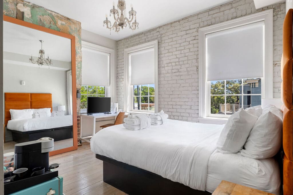 Bright hotel-style bedroom with white bedding, an orange headboard seen in the mirror, exposed brick wall, and a chandelier.