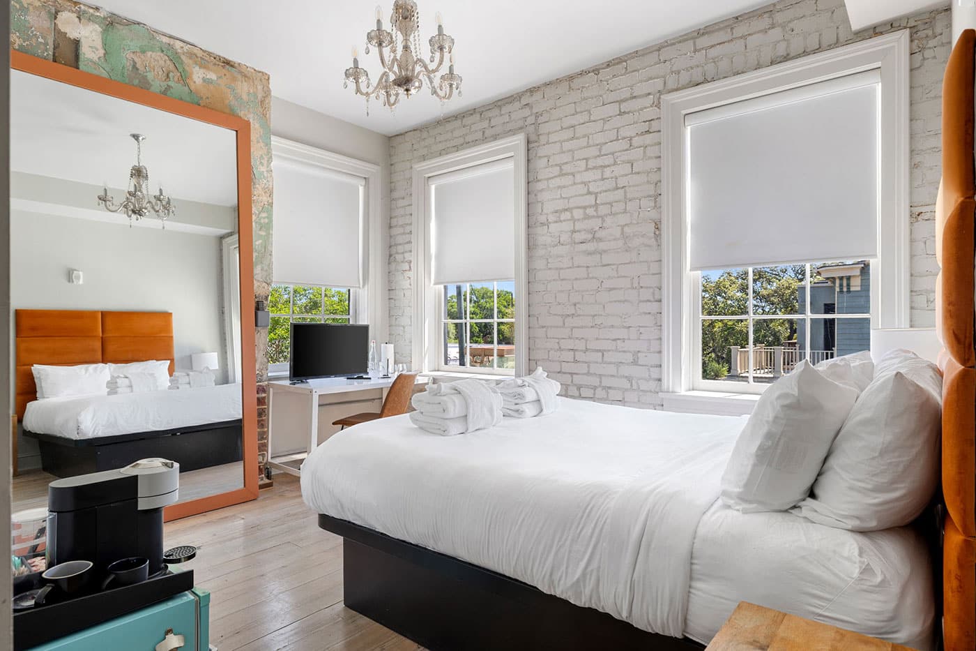 Bright hotel-style bedroom with white bedding, an orange headboard seen in the mirror, exposed brick wall, and a chandelier.