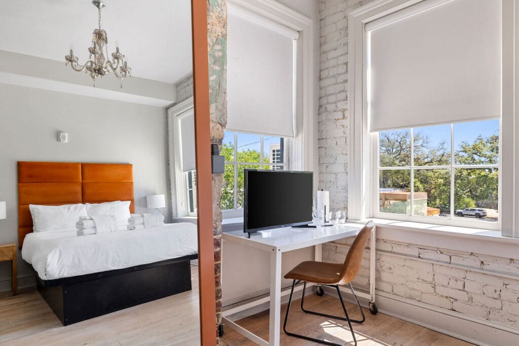 Loft-style bedroom with white bedding and an orange padded headboard, chandelier, and a white desk by large windows.
