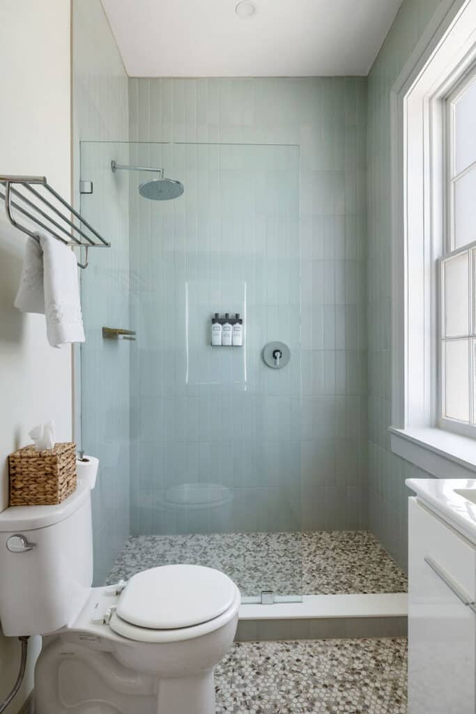 Walk-in shower with light blue vertical tiles and a glass enclosure, rainfall showerhead, three shampoo dispensers, and a nearby white toilet with a tissue box on top of the tank; pebble tile floor throughout the room.