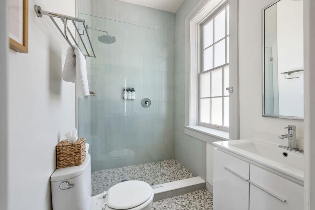 Bright modern bathroom with a glass-enclosed shower, pale blue tile walls, and a white vanity against a windowed wall.