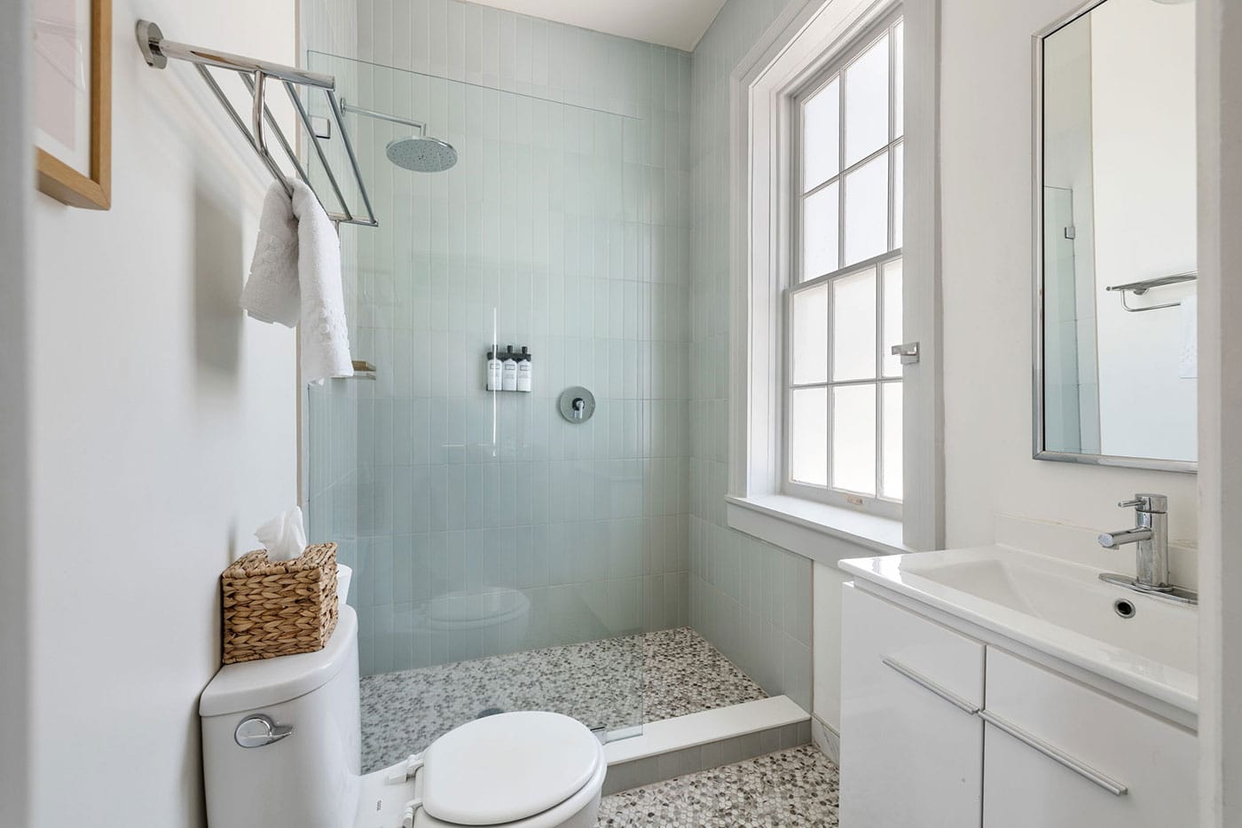 Bright modern bathroom with a glass-enclosed shower, pale blue tile walls, and a white vanity against a windowed wall.