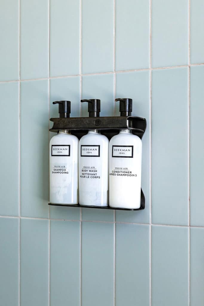 Three white pump bottles in a black wall-mounted rack against pale blue tiled shower wall, labeled shampoo, body wash, and conditioner by Beekman.