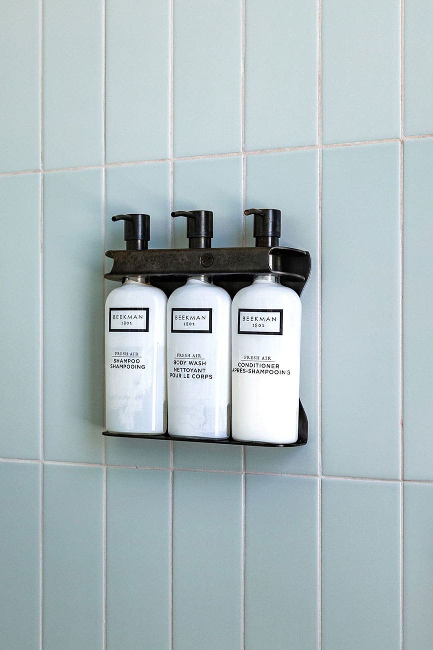 Three white pump bottles in a black wall-mounted rack against pale blue tiled shower wall, labeled shampoo, body wash, and conditioner by Beekman.