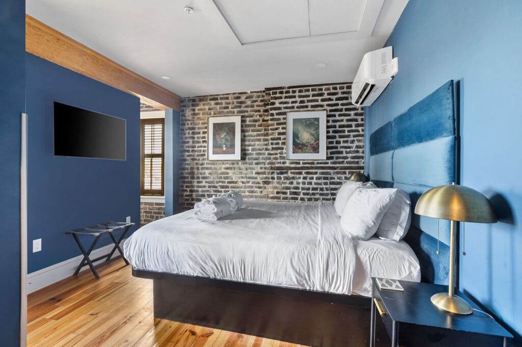 Blue-toned bedroom with a white bed, brick accent wall, two framed pictures, and a wall-mounted TV on the blue wall at left.